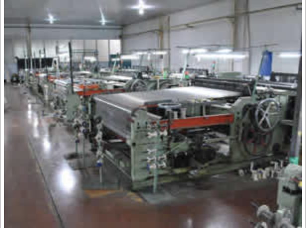 A row of shuttle looms weaving wire mesh inside a lit production hall